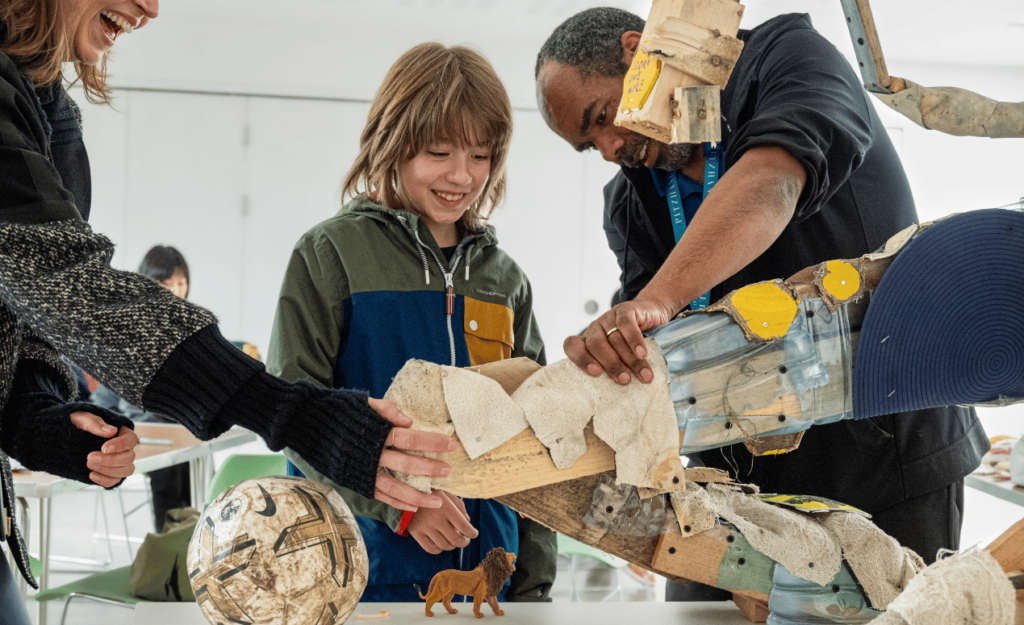 Children and a facilitator work together, adding elements to a lion sculpture during a community art workshop.